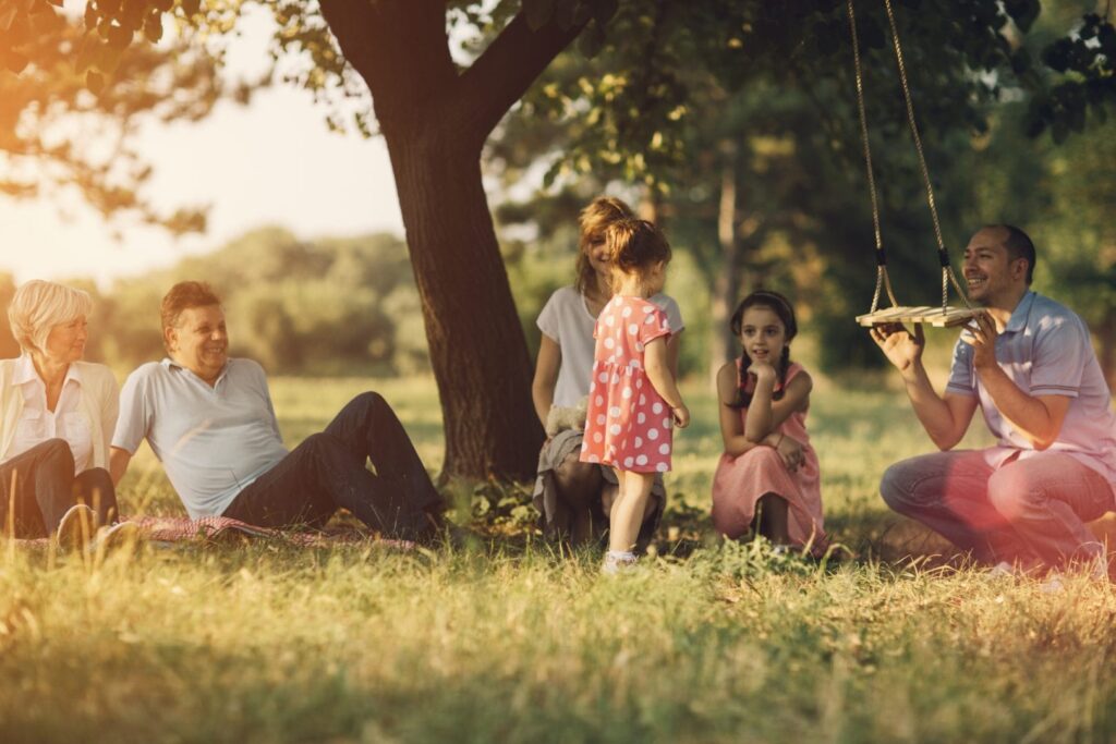 A group of adults sitting on a lawn with young children, playing with a swing hanging from a tree