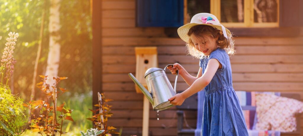 A young girl in a dress and a sun hat using a watering can on shrubs